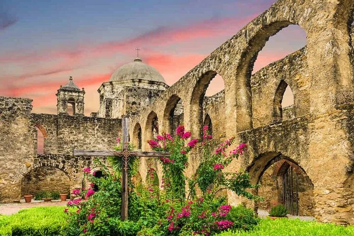 Stone arches of a mission ruin with pink flowers and a cross